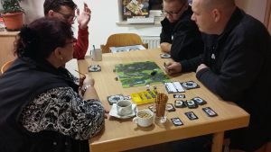Bild: Tisch mit Spielmaterial, an dem 4 erwachsene Spieler sitzen