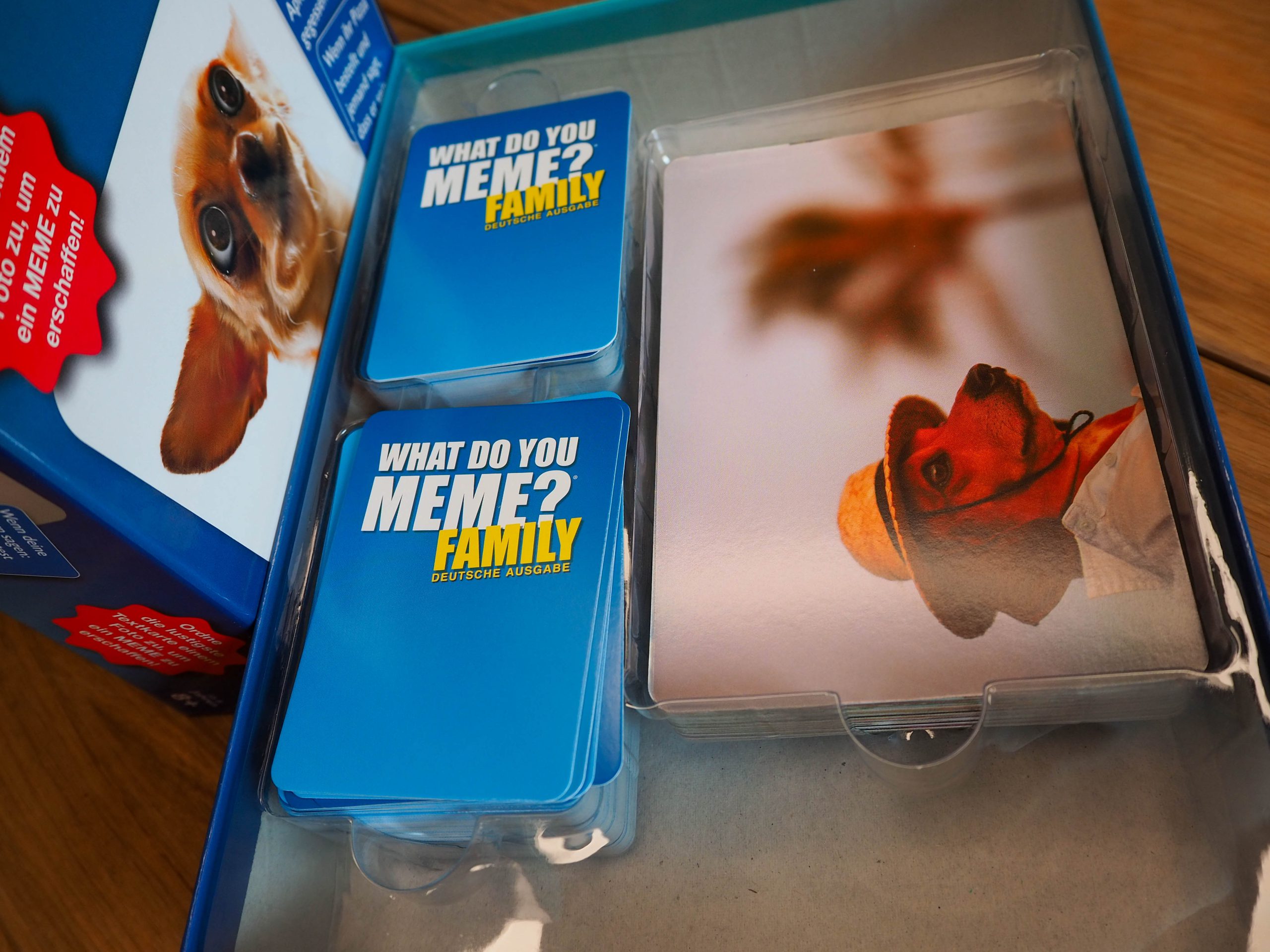 Die offene Spielschachten liegt im Bild. Darin zu sehen eine dominante Plastikeinlage mit zwei Stapeln Textkarten und einem Stapel Bildkarten. Links neben der offenen Spielschachtel steht hochkant der Deckel.