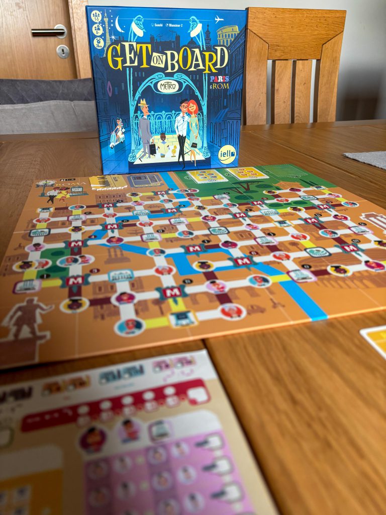 Die Spielschachtel steht aufrecht auf dem Tisch. Davor liegt das Spielplan und direkt davor der Spielerblock. Dieser ist nur verschwommen zu sehen.
