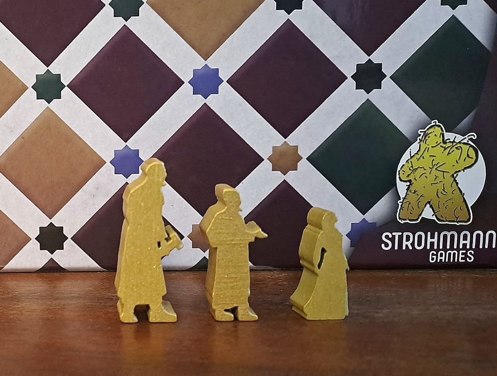 Zu sehen sind die unterschiedlichen Spielfiguren vor der Spielschachtel. Baumeister, Händler und Dichterin.