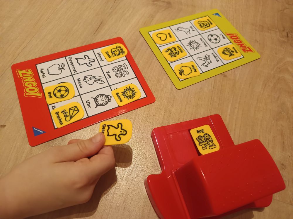 Auf dem Bild liegt ein Beispiel des Spiels für zwei Personen vor. Einige Kärtchen sind bereits gelegt. Der Begriff Gespenst wurde gerade genannt und der Spieler hält ihn in der Hand, um ihn auf der Legetafel zu platzieren.