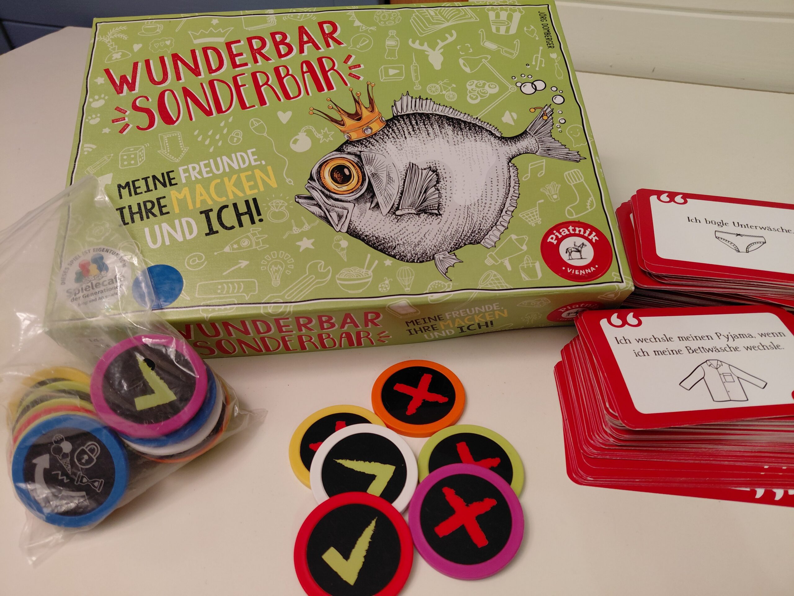 Auf dem Tisch liegen die Verpackung, verschiedene Spielchips mit grünem Haken oder rotem Kreuz und ein Stapel mit Aufgabenkarten.